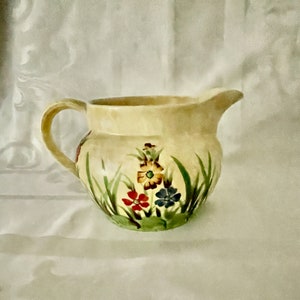 May include: A cream-coloured ceramic jug with a floral design. The jug has a wide mouth and a handle. The flowers are painted in shades of red, yellow, blue, and green.