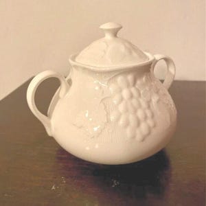 May include: A white ceramic sugar bowl with a matching lid, featuring two side handles and a raised, embossed pattern of grape clusters and vine leaves on the exterior surface. The bowl has a rounded body and a smooth, glossy finish.