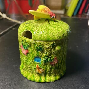 May include: A green ceramic jar with a lid, featuring a yellow and red insect on top. The jar is adorned with floral and insect designs. A small opening is on the side of the lid. The jar is decorated with floral and insect motifs.