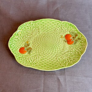 May include: A green ceramic serving dish with a woven basket design and two red tomato accents.
