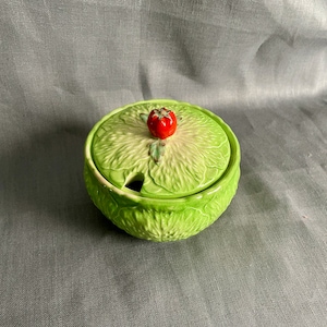 May include: A green ceramic sugar bowl with a lid shaped like a lettuce leaf. The lid has a small red strawberry on top.