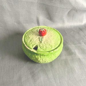 May include: A green ceramic jar and lid, shaped like a cabbage, with a red tomato on top. The jar has a small opening for a spoon. The jar is on a grey surface.