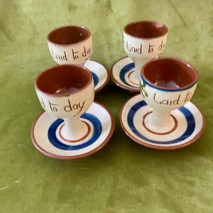 May include: Four ceramic egg cups with matching saucers. The egg cups are cream-coloured with brown interiors and the words "Laid to day" in brown script. The saucers have blue and brown concentric circles.