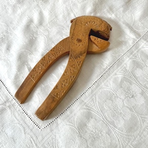 May include: A vintage wooden nutcracker with detailed carvings. The light brown nutcracker has a lever design with decorative patterns on the handles and head. It rests on a white tablecloth with a floral pattern. The nutcracker is likely from the mid-20th century.