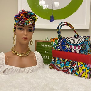 May include: A mannequin head wearing a colorful head wrap, gold chain necklace, and earrings. A patterned tote bag with red accents sits next to the mannequin. The bag and head wrap share a similar vibrant design.