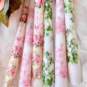 May include: Six white taper candles with floral designs in pink and green. The candles feature roses, greenery, and decorative elements. Some candles have gold accents. The candles are arranged on a white, textured surface.