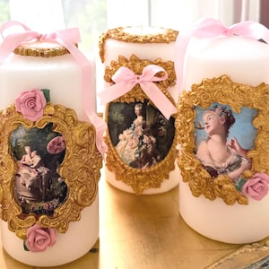 Könnte beinhalten: Weiße Säulenkerzen mit goldenen Rahmen mit klassischen Kunstdrucken. Jede Kerze ist mit einem rosa Band und einer rosa Rose verziert. Die Kerzen sind dekorativ und elegant.