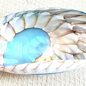 May include: A decorative, oval-shaped glass bowl with a unique design. The bowl features a light blue center and a textured, shell-like pattern in shades of white, gray, and brown. The rim is a darker shade of blue.