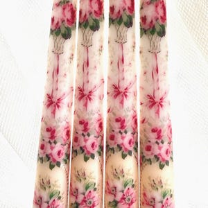 May include: Four ivory taper candles with a floral design. The candles feature a repeating pattern of pink roses, green leaves, and gold accents.