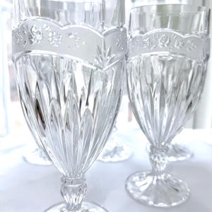 May include: Clear crystal wine glasses with a decorative floral band near the rim. The glasses have a detailed, cut-glass design on the bowl and a pedestal base. The background is a soft, out-of-focus white.