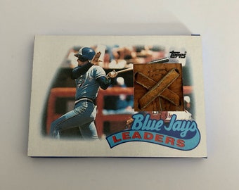 May include: A baseball card featuring a baseball player in a blue uniform swinging a bat. The card has a brown leather baseball glove patch with the text "Blue Jays Leaders" in blue and white.