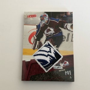 May include: A hockey card featuring Patrick Roy, a former NHL goalie. The card is from the Victory series and features a blue and white patch of the Colorado Avalanche logo.