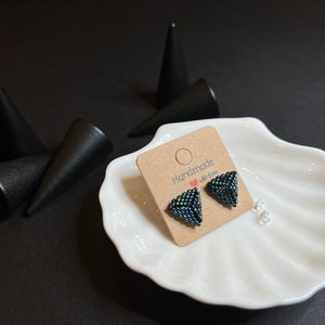 May include: A pair of handmade triangle-shaped earrings with a dark blue and green iridescent finish. The earrings are displayed on a white dish with a brown card that reads "Handmade with Love".