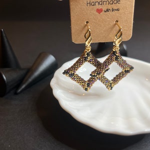 Könnte beinhalten: Ein Paar goldfarbene Ohrringe mit einem geometrischen Muster in Schwarz und Lila. Die Ohrringe werden auf einer weißen, muschelförmigen Schale präsentiert. Das Etikett lautet "Handmade with love".
