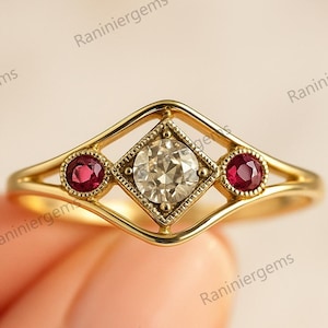 Może przedstawiać: Złoty pierścionek z centralnym kamieniem przypominającym diament w kwadratowej oprawie, otoczonym dwoma mniejszymi, okrągłymi czerwonymi kamieniami. Pierścionek ma ażurowy wzór z delikatnym, zakrzywionym paskiem.
