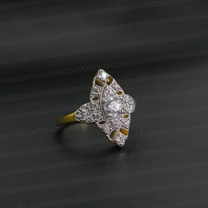 May include: A marquise-shaped ring featuring a central round stone surrounded by smaller pavé-set stones in a silver-toned metal setting. The band is gold-toned, and the intricate head of the ring displays openwork filigree details throughout.