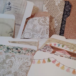May include: A collection of decorative file folders with various patterns. Some folders feature floral, lace, and vintage-style designs. The color palette includes cream, beige, pink, and touches of other colors. The folders have a tabbed design for easy organization.