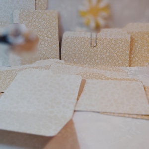 May include: A collection of stationery items, including envelopes and cards, arranged on a white surface. The items feature a cream-colored floral pattern. Some envelopes are secured with a silver paper clip.