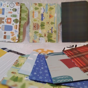 May include: A collection of decorative file folders and patterned papers. The folders feature camping and outdoor-themed designs with images of a bear, trees, and camping gear. The papers have various patterns, including plaid, stars, and geometric shapes.
