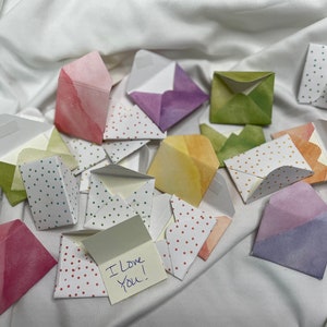 May include: A collection of small, colorful envelopes with polka dot patterns. The envelopes are made of paper and have a watercolor effect. One envelope is open and has a note inside that says "I love you!"