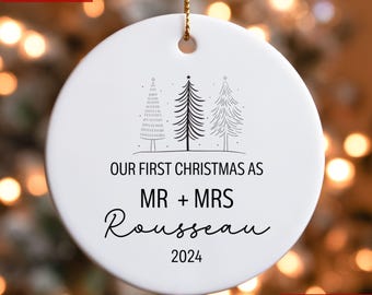 Könnte beinhalten: Weißer Weihnachtsbaumschmuck aus Keramik mit schwarzem Text, der "Unser erstes Weihnachten als Herr und Frau Rousseau 2024" lautet. Der Schmuck zeigt eine einfache Linienzeichnung von drei Weihnachtsbäumen.