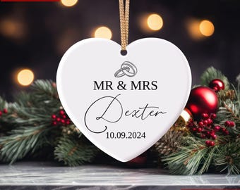 Könnte beinhalten: Ein weißer, herzförmiger Weihnachtsschmuck mit goldenem Band, mit dem Text "MR & MRS Dexter" und dem Datum "10.09.2024". Der Schmuck hat eine Grafik von Eheringen und ist von festlichem Grün und roten Ornamenten umgeben.