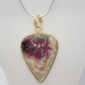 May include: A teardrop-shaped pendant with a gold-colored bezel and bail, suspended from a silver chain. The pendant features a polished stone with a mix of colors, including pink, green, and cream. The stone has a unique, natural pattern.
