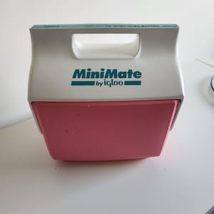 May include: A white and pink MiniMate cooler by Igloo. The cooler has a white handle and a pink lower section. The text "MiniMate by Igloo" is printed in teal on the front. Designed to keep items cold, it's ideal for picnics or days out.