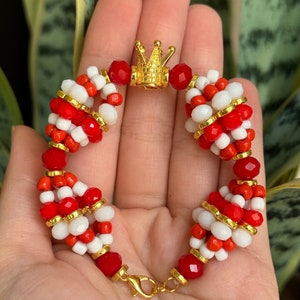May include: A red and white beaded bracelet with a gold crown charm. The bracelet is made with small, round beads and has a clasp closure.