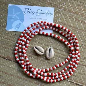 May include: Red and white beaded waist beads with two cowrie shells. The beads are handmade and the card in the background says "Ekes Olomidara Handmade with Love".