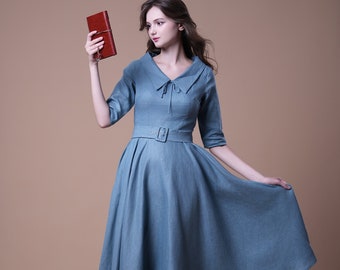 以下が含まれることがあります： 白い襟と茶色のベルトが付いた薄い青色のドレスを着た女性。彼女は右手に小さな茶色の本を持っています。