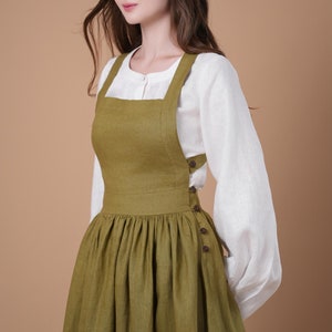 May include: A woman wearing a green linen overall dress with a white blouse. The dress has a square neckline and a gathered skirt. The dress is fastened with buttons on the side.