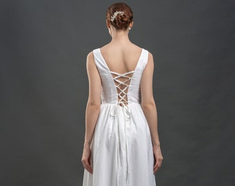 May include: A white sleeveless dress with a corset-style back and a flowing skirt. The dress features a lace-up detail on the back and a tie at the waist. The model is wearing black heels. The dress is suitable for special occasions.