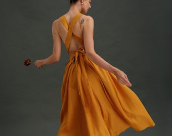 May include: A woman wearing a mustard yellow sundress with a criss-cross back detail. The dress has a flowy skirt and is cinched at the waist with a tie. She is wearing black heeled sandals and holding a single red rose.