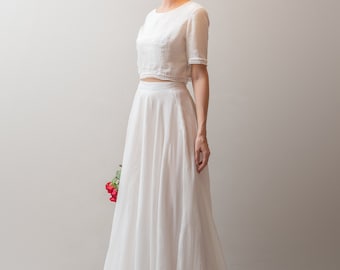 May include: A white two-piece bridal outfit with a cropped top and a flowing skirt. The top has a lace trim and the skirt is made of a lightweight fabric.