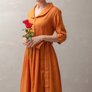 Puede incluir: Una mujer lleva un vestido largo de lino color &oacute;xido con cuello blanco y cintur&oacute;n a juego. Lleva un ramo de rosas rojas.