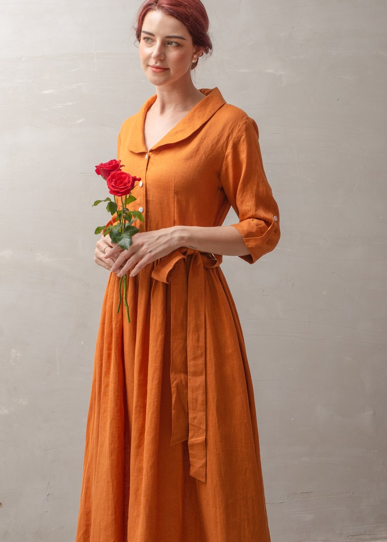 Puede incluir: Una mujer lleva un vestido largo de lino color &oacute;xido con cuello blanco y cintur&oacute;n a juego. Lleva un ramo de rosas rojas.