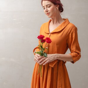 Puede incluir: Una mujer con un vestido de lino naranja quemado sostiene un ramo de tres rosas rojas. El vestido tiene un cuello y un cintur&oacute;n de lazo en la cintura.