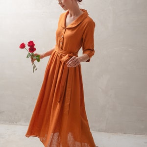 Puede incluir: Una mujer con un vestido largo de lino color &oacute;xido con cuello blanco y cintur&oacute;n a juego. Lleva un ramo de rosas rojas y las mira.