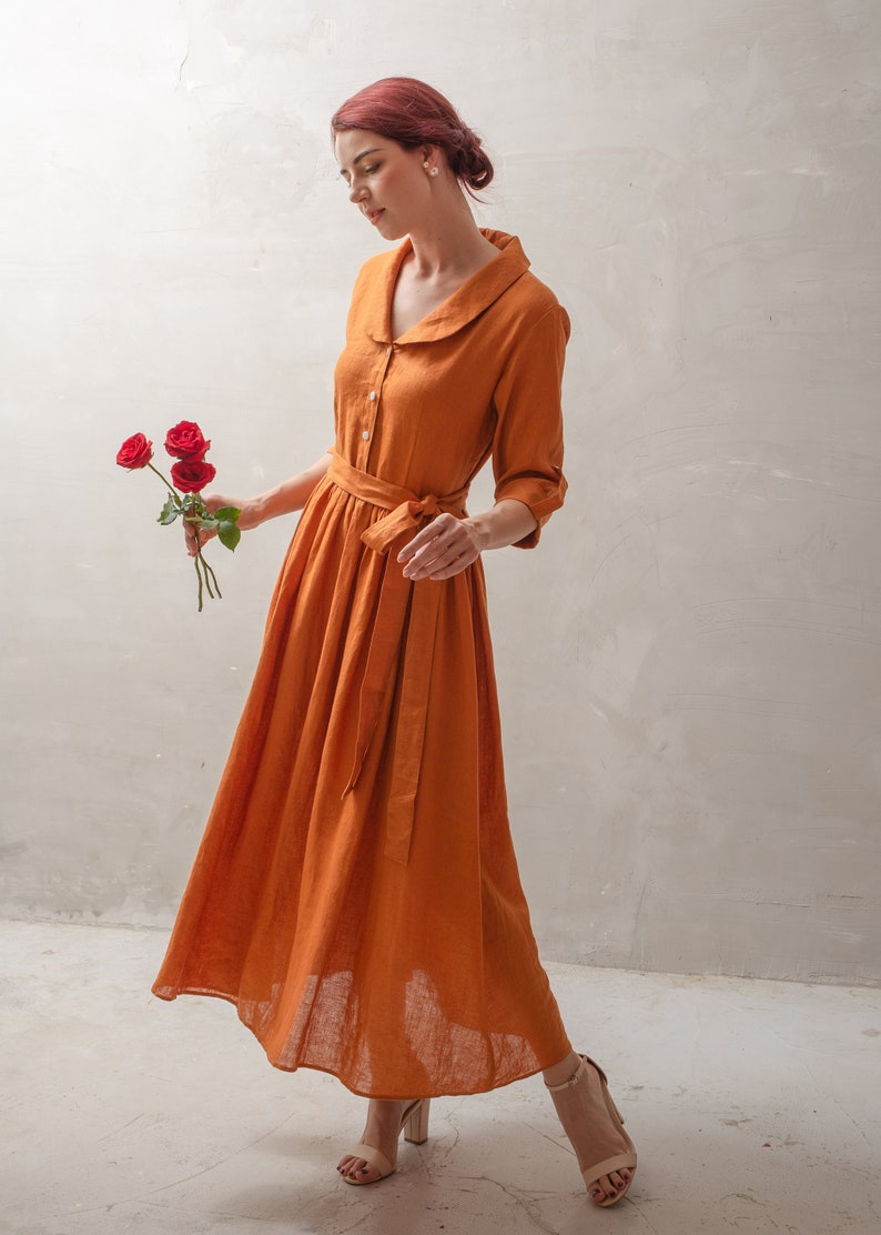 Puede incluir: Una mujer con un vestido largo de lino color &oacute;xido con cuello blanco y cintur&oacute;n a juego. Lleva un ramo de rosas rojas y las mira.
