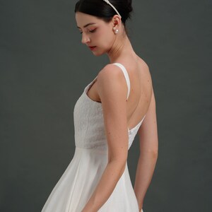 May include: A white sleeveless wedding dress with a fitted bodice and a flowing skirt. The dress has a simple design with a scoop neckline and a low back. The model is wearing a pearl headband and pearl earrings.