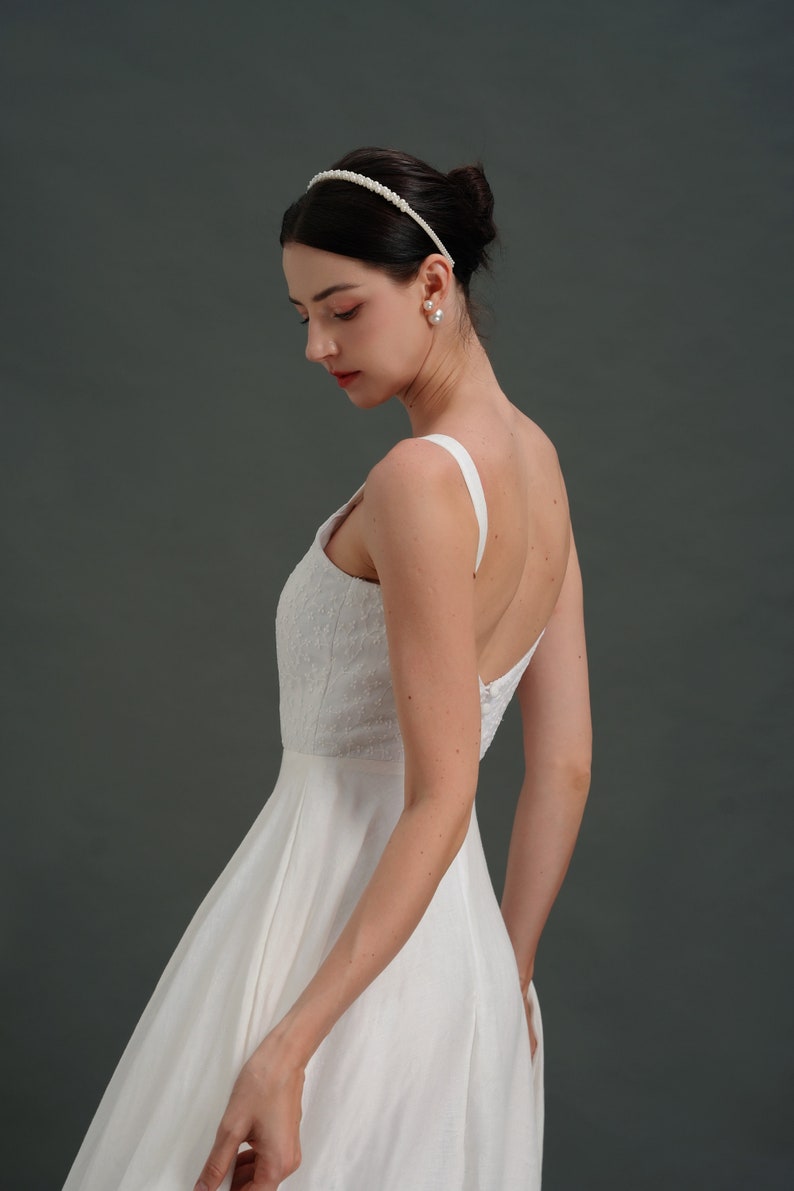 May include: A white sleeveless wedding dress with a fitted bodice and a flowing skirt. The dress has a simple design with a scoop neckline and a low back. The model is wearing a pearl headband and pearl earrings.