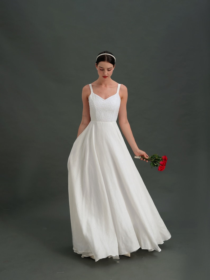 May include: A white sleeveless wedding dress with a sweetheart neckline and a flowing skirt. The bride is holding a bouquet of red roses.