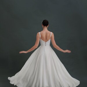 May include: A white wedding dress with a simple, flowing skirt and a sleeveless bodice. The dress has a low back and a zipper closure.