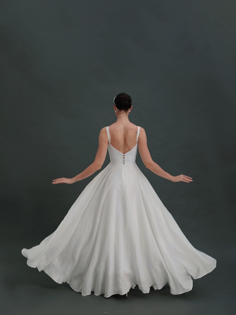 May include: A white wedding dress with a simple, flowing skirt and a sleeveless bodice. The dress has a low back and a zipper closure.