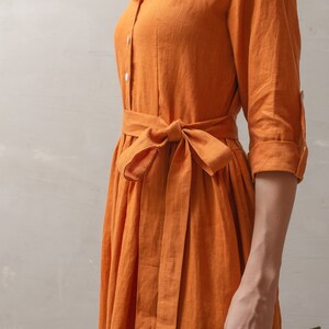 Puede incluir: Una mujer lleva un vestido largo de lino naranja con un cintur&oacute;n de lazo. El vestido tiene un cuello y mangas largas. Lleva un ramo de rosas rojas.