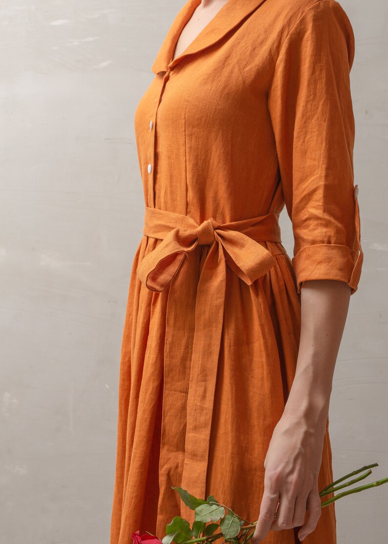 Puede incluir: Una mujer lleva un vestido largo de lino naranja con un cintur&oacute;n de lazo. El vestido tiene un cuello y mangas largas. Lleva un ramo de rosas rojas.