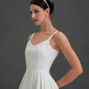 May include: A white sleeveless dress with a fitted bodice and a full skirt. The dress has a delicate floral pattern and a simple v-neckline. The model is wearing a pearl headband and pearl earrings.