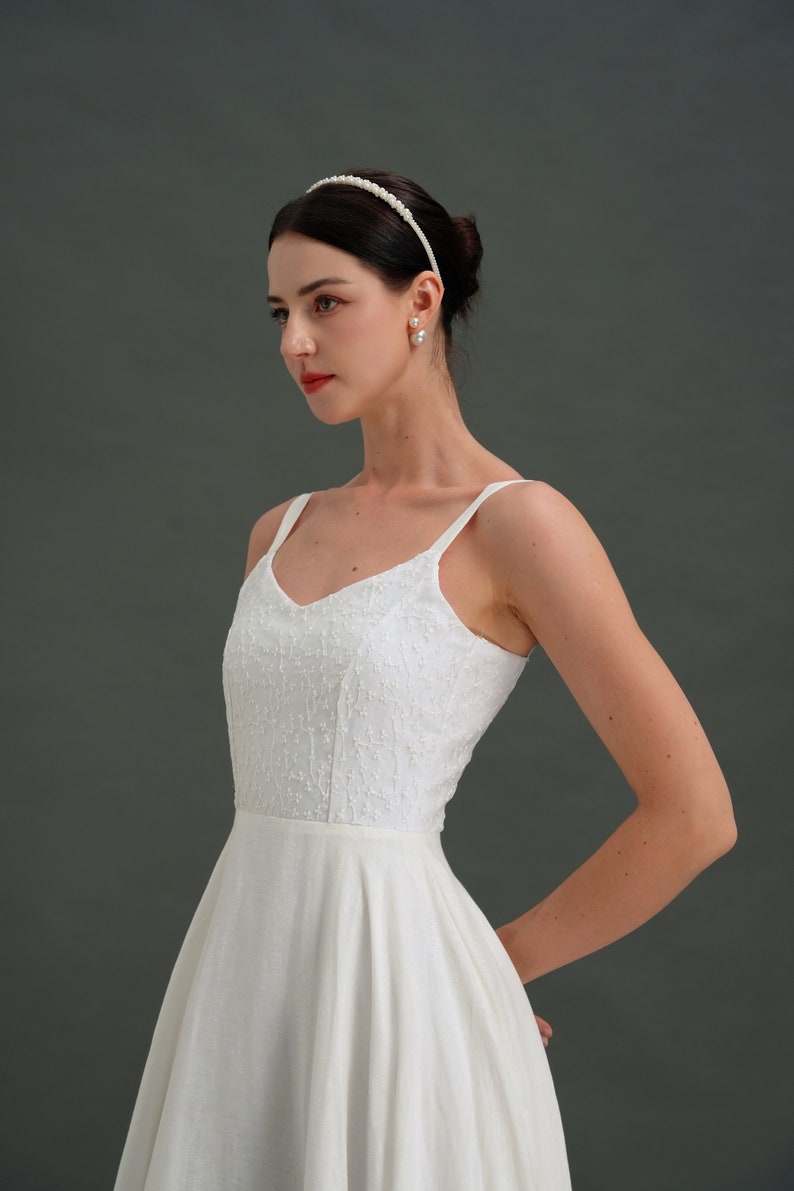May include: A white sleeveless dress with a fitted bodice and a full skirt. The dress has a delicate floral pattern and a simple v-neckline. The model is wearing a pearl headband and pearl earrings.