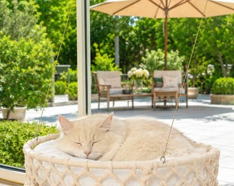 May include: A cream-colored cat sleeping in a round, macrame-style hanging window bed. The bed features a woven rope lattice frame with a soft white cushion inside, suspended by thin wires attached to the window with clear suction cups.